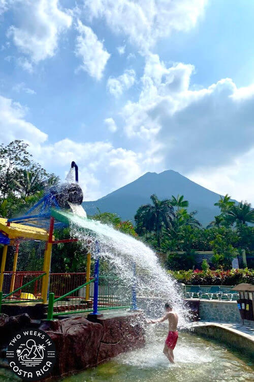 a kid at a water play structure with a volcano in the background