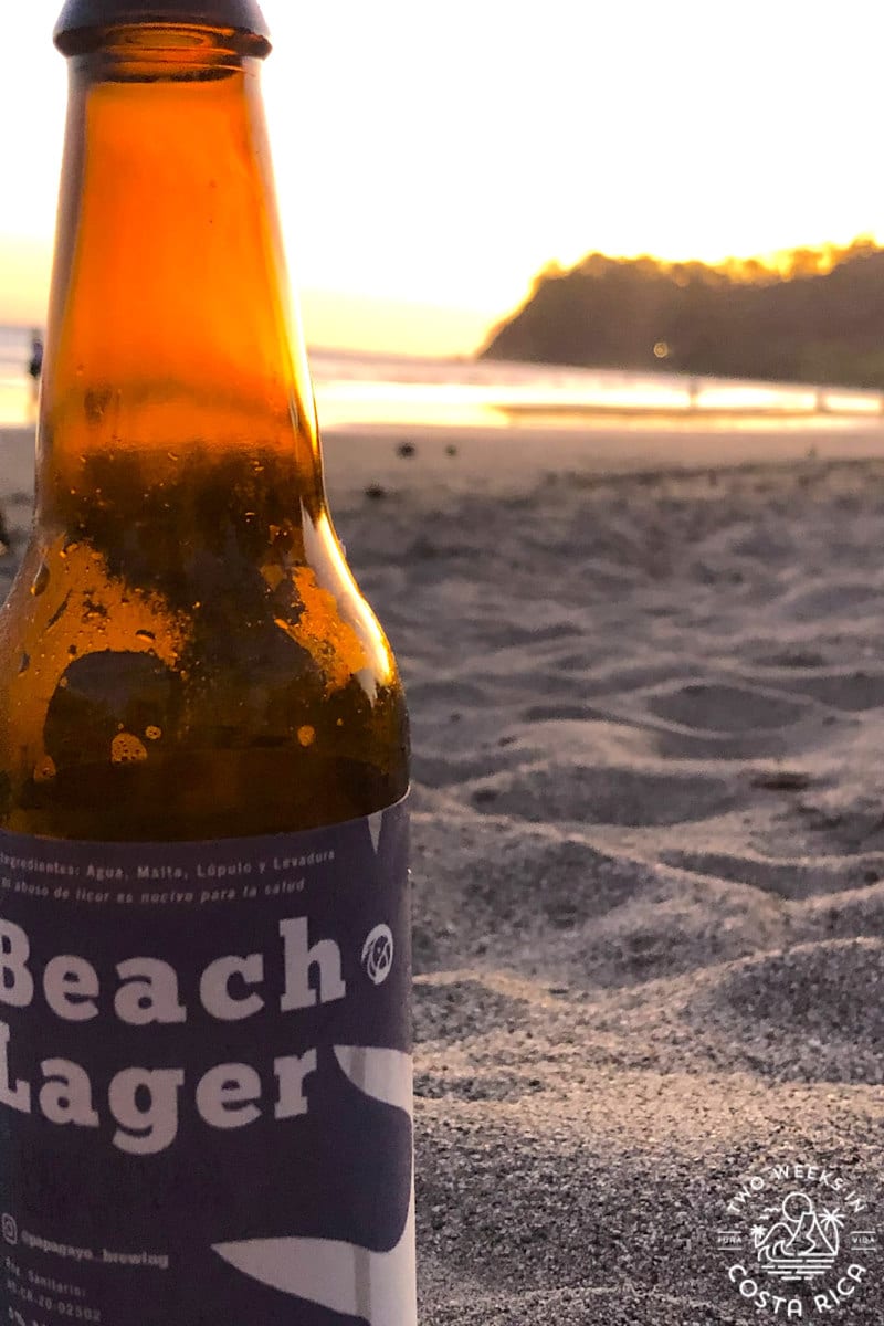 A bottle of beer on the sand with an ocean sunset in the background