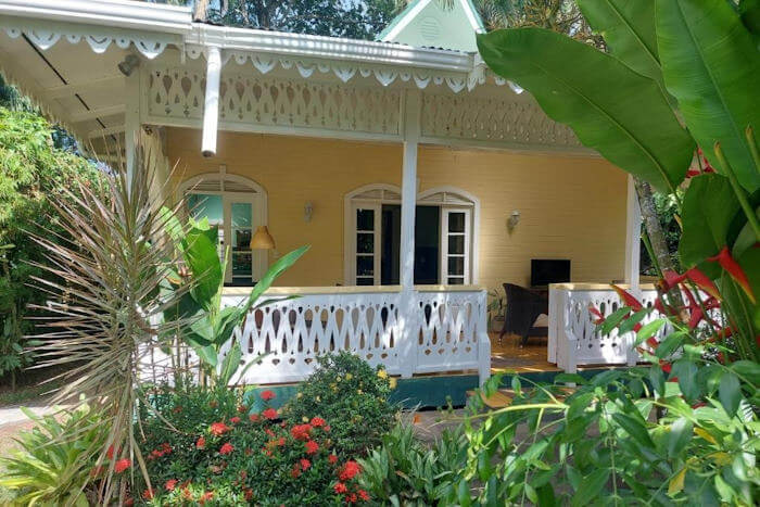 A yellow house with intricate white trip and a porch, set in a tropical garden. 