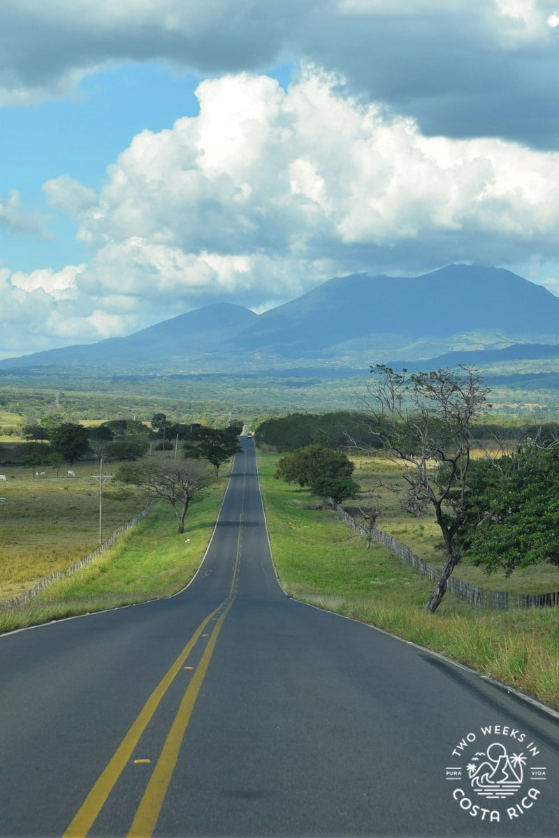 a straight road with grass on both sides and steep mountains in the distance